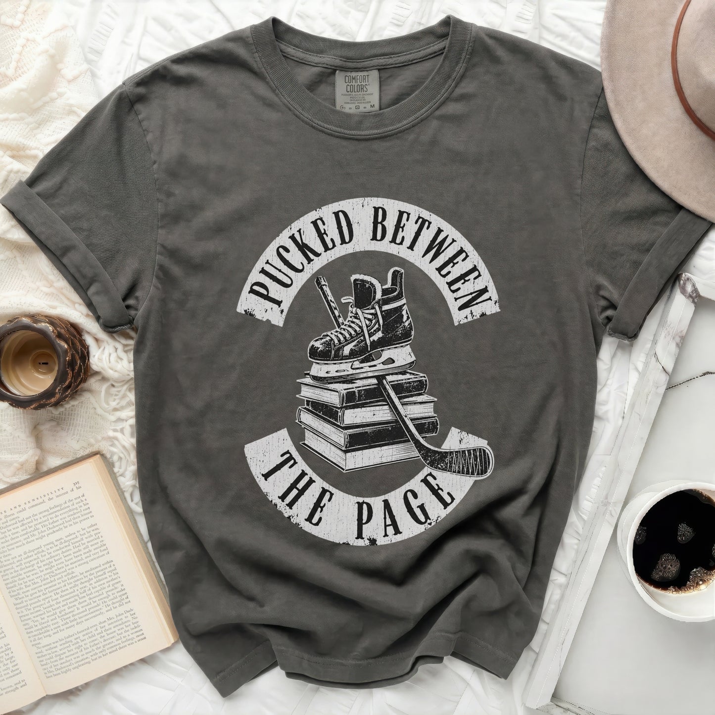 Dark grey t-shirt with white hockey skates, stick, and text "Pucked Between The Page".