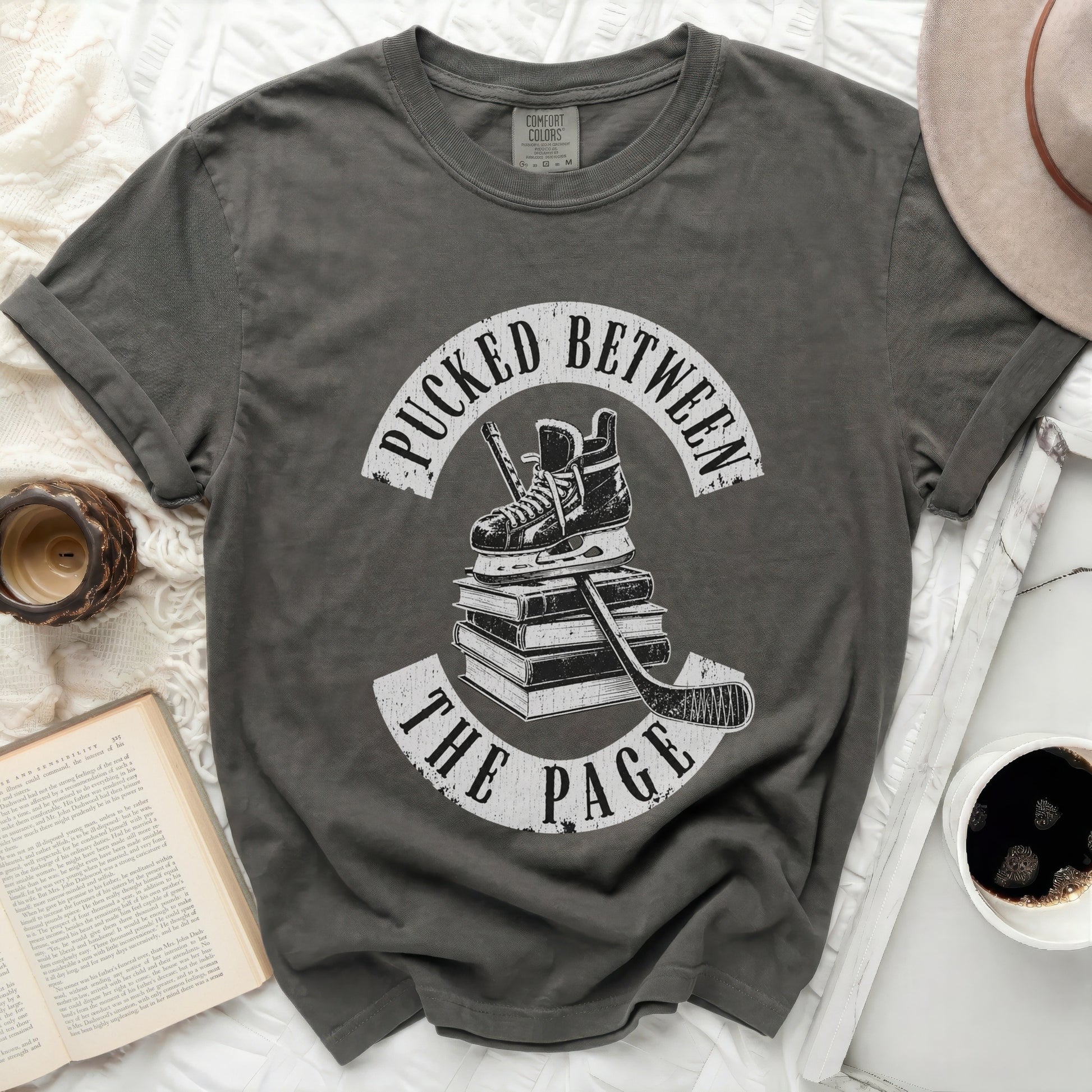 Dark grey t-shirt with white hockey skates, stick, and text "Pucked Between The Page".