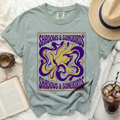 Green t-shirt with gold and purple "Shadows & Songbirds" graphic.