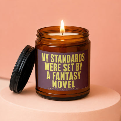 Amber glass candle with lit wick and purple label reading "MY STANDARDS WERE SET BY A FANTASY NOVEL".