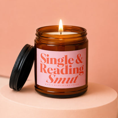 Amber glass candle with lit flame and pink label reading "Single & Reading Smut".