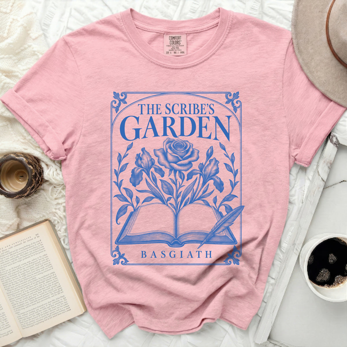Pink t-shirt with blue graphic of an open book, rose, iris, and feather. Text: THE SCRIBE'S GARDEN BASGIATH.