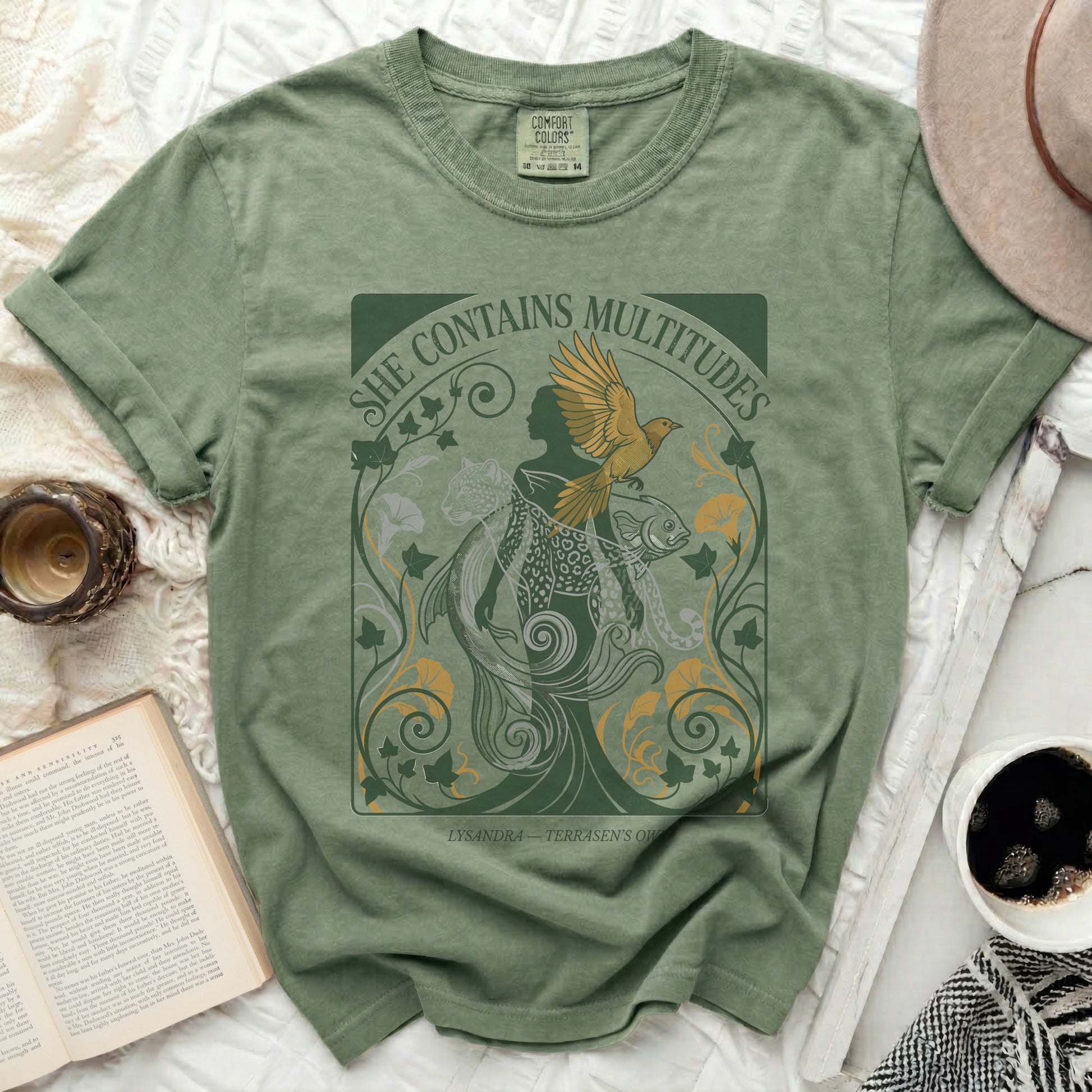 Olive green t-shirt with gold "She Contains Multitudes" text and mythical graphic.