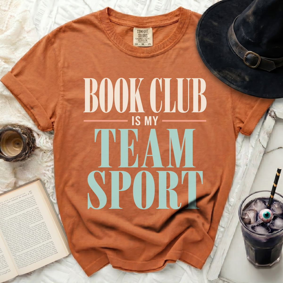 Orange t-shirt with "Book Club Is My Team Sport" text, next to a hat and book.