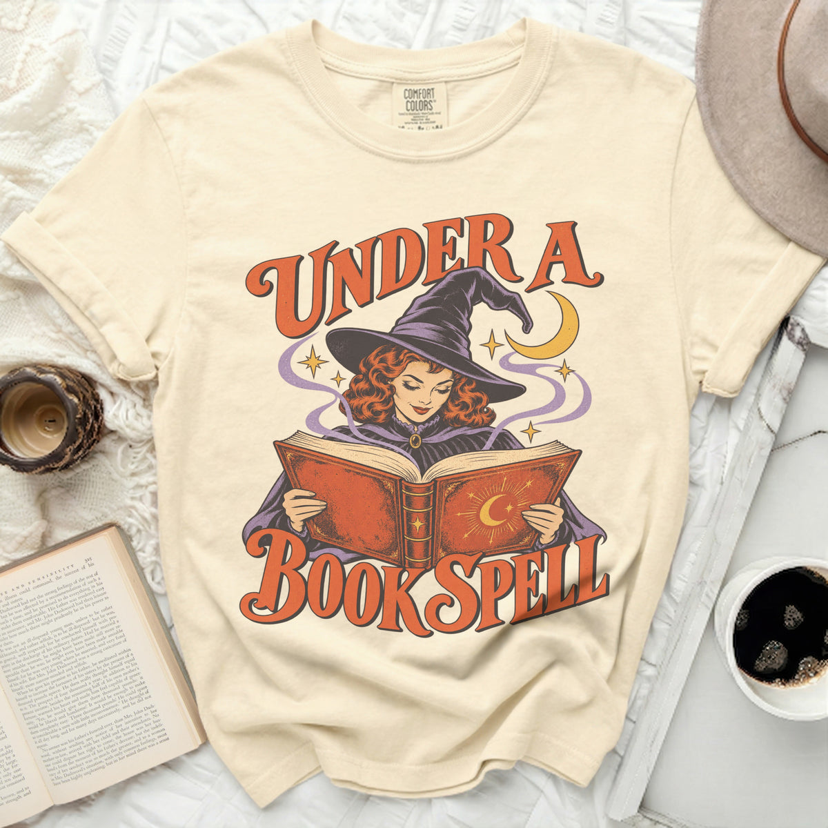 Cream t-shirt with a witch reading a book, text reads "Under a Book Spell".