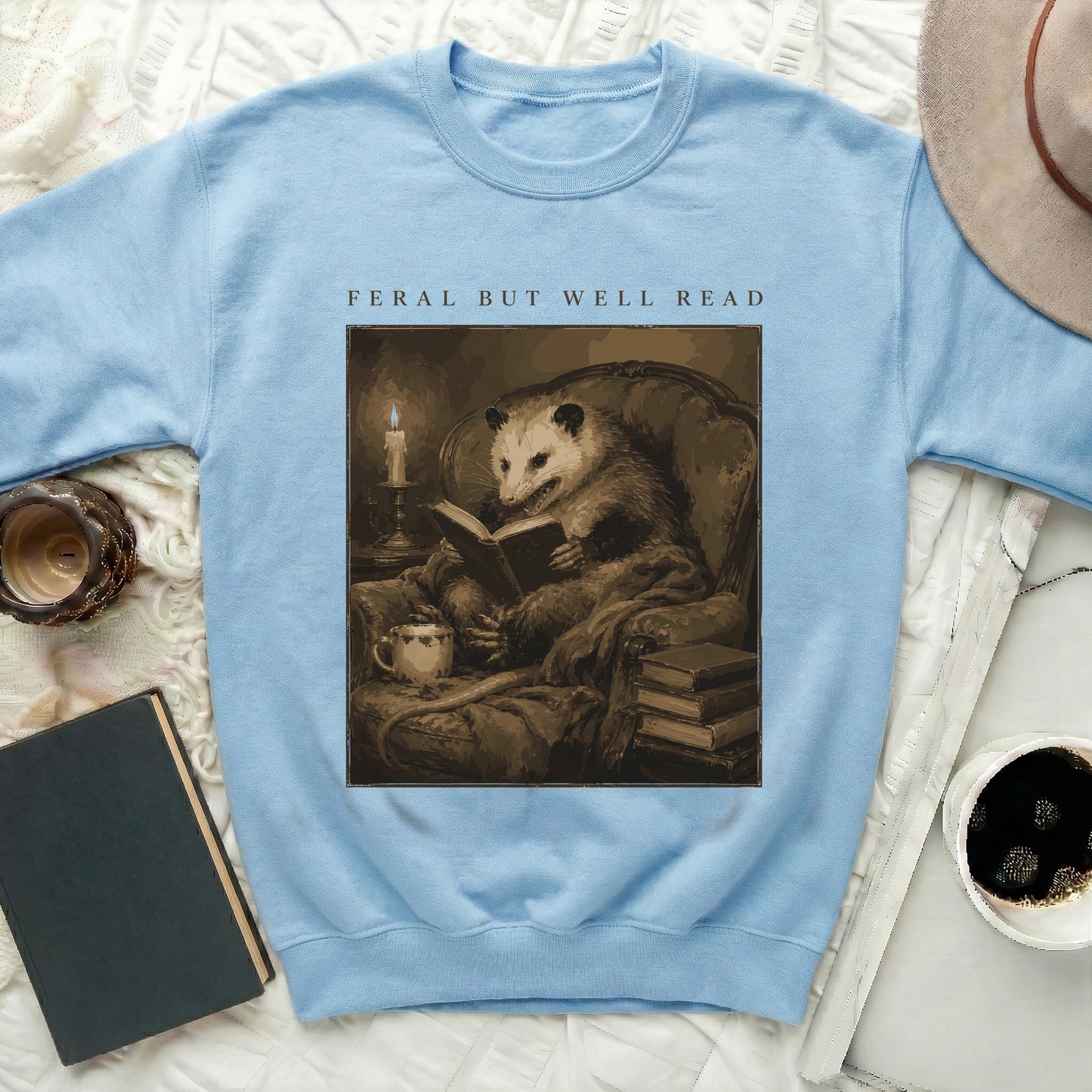 Light blue sweatshirt featuring an opossum reading on a chair, text "FERAL BUT WELL READ."