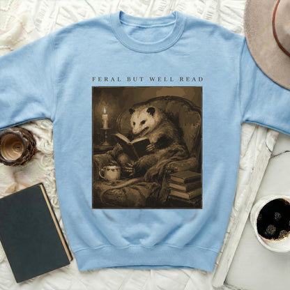 Light blue sweatshirt featuring an opossum reading on a chair, text "FERAL BUT WELL READ."