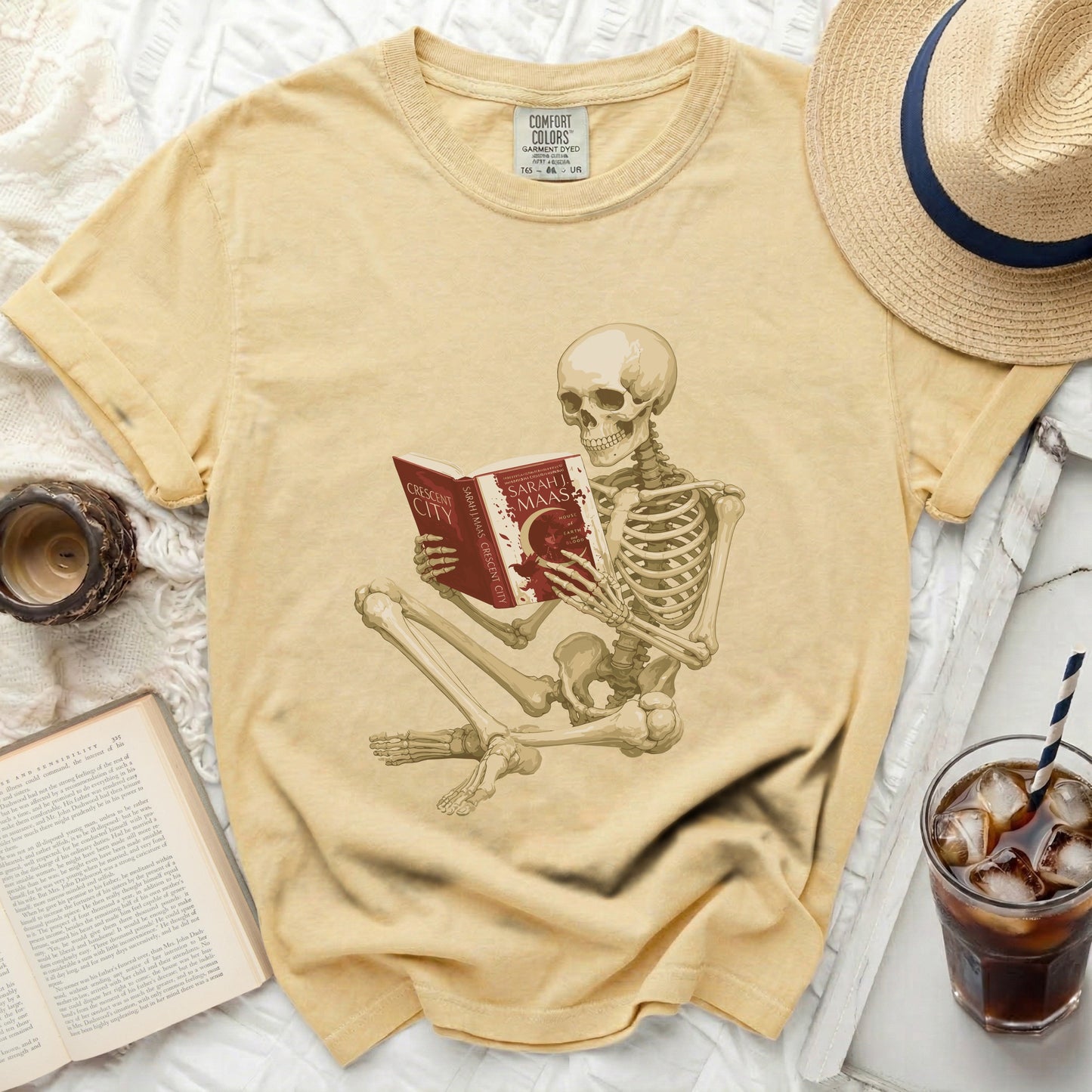 Skeleton reading a red book titled "Crescent City" by Sarah J. Maas, on a yellow shirt.