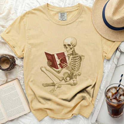 Skeleton reading a red book titled "Crescent City" by Sarah J. Maas, on a yellow shirt.