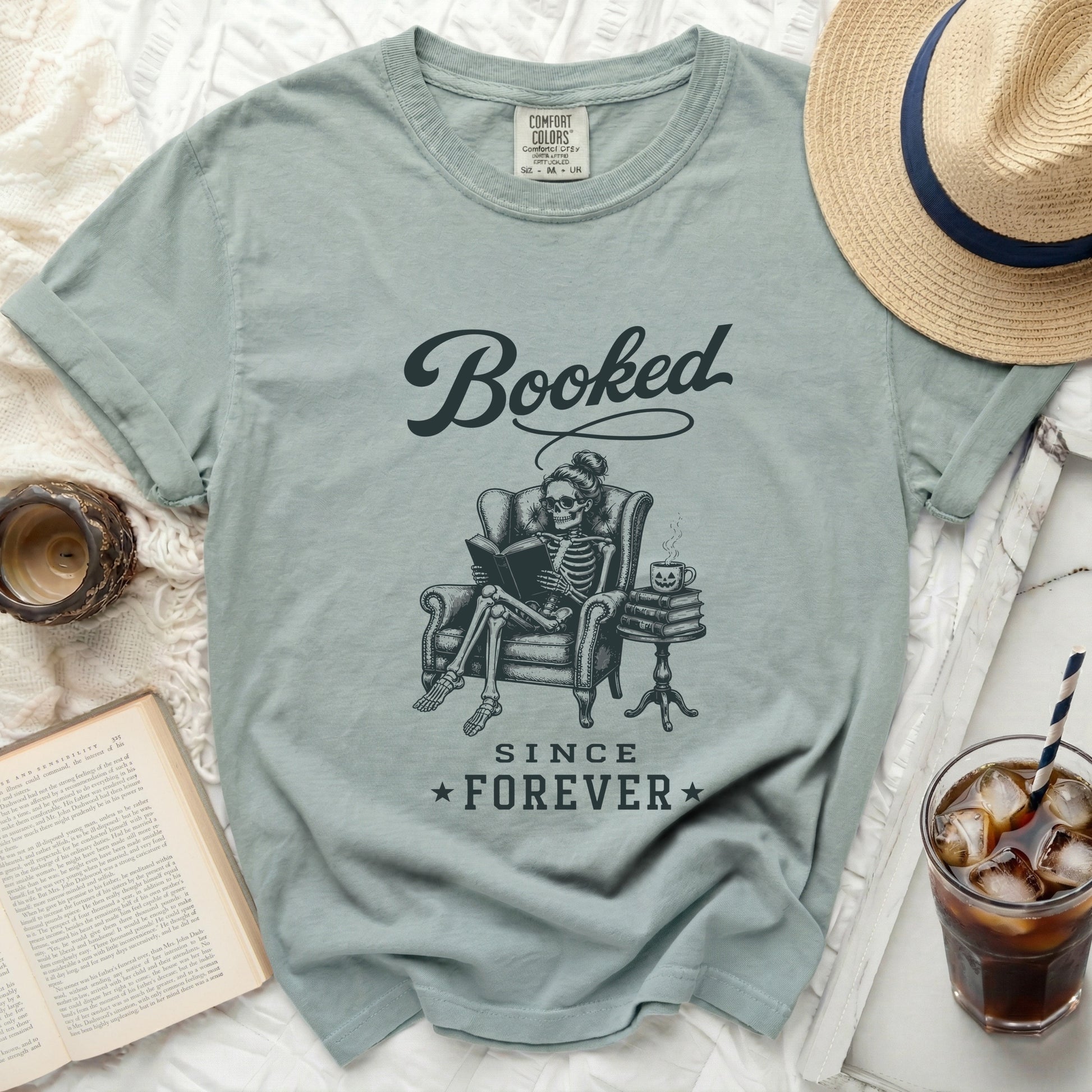 Pale green t-shirt featuring a skeleton reading a book, titled "Booked Since Forever".