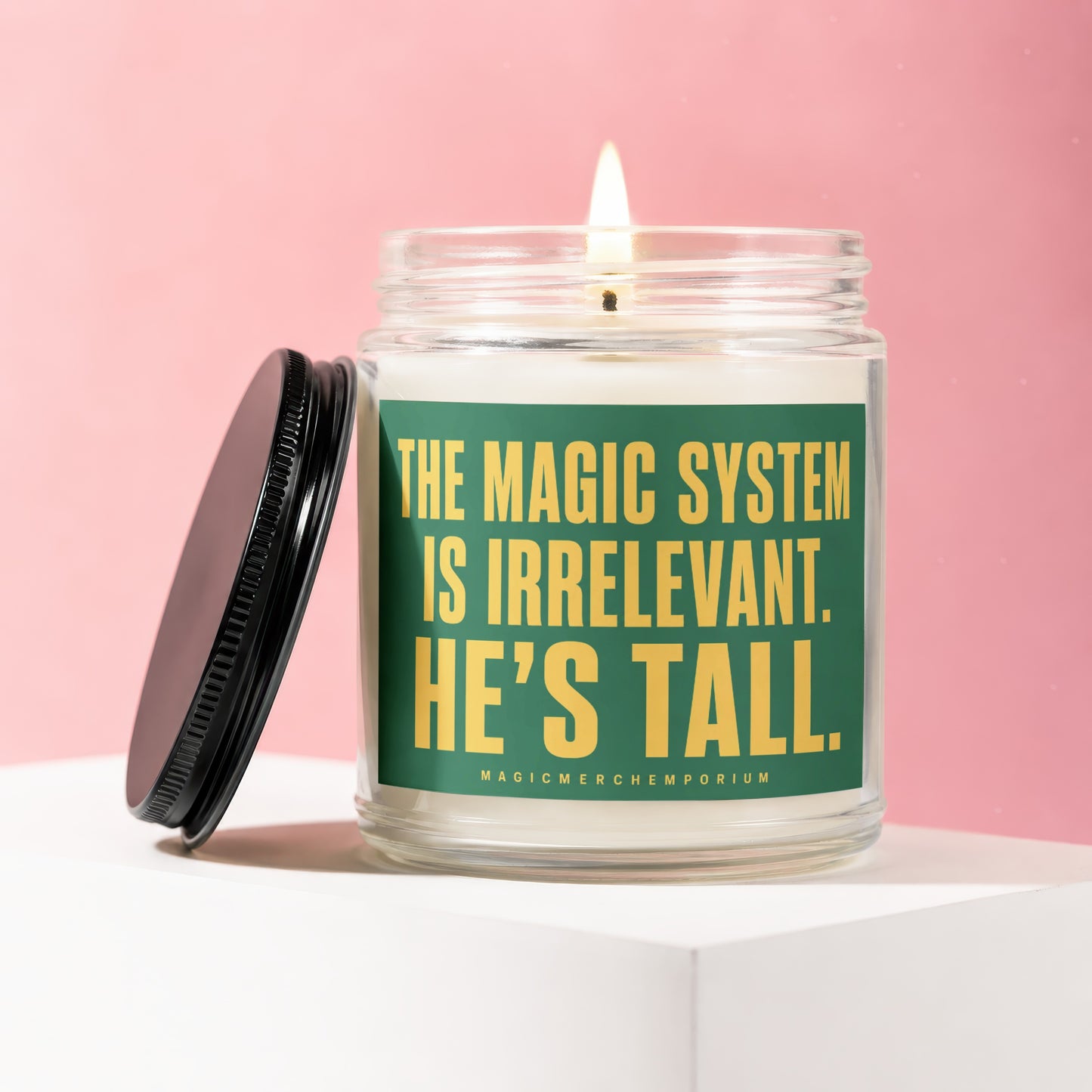 Glass candle with lit wick and black lid, featuring text "THE MAGIC SYSTEM IS IRRELEVANT. HE'S TALL."
