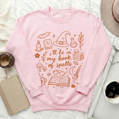 Pink crewneck sweatshirt with "I'll be in my book of spells" and witchy illustrations.