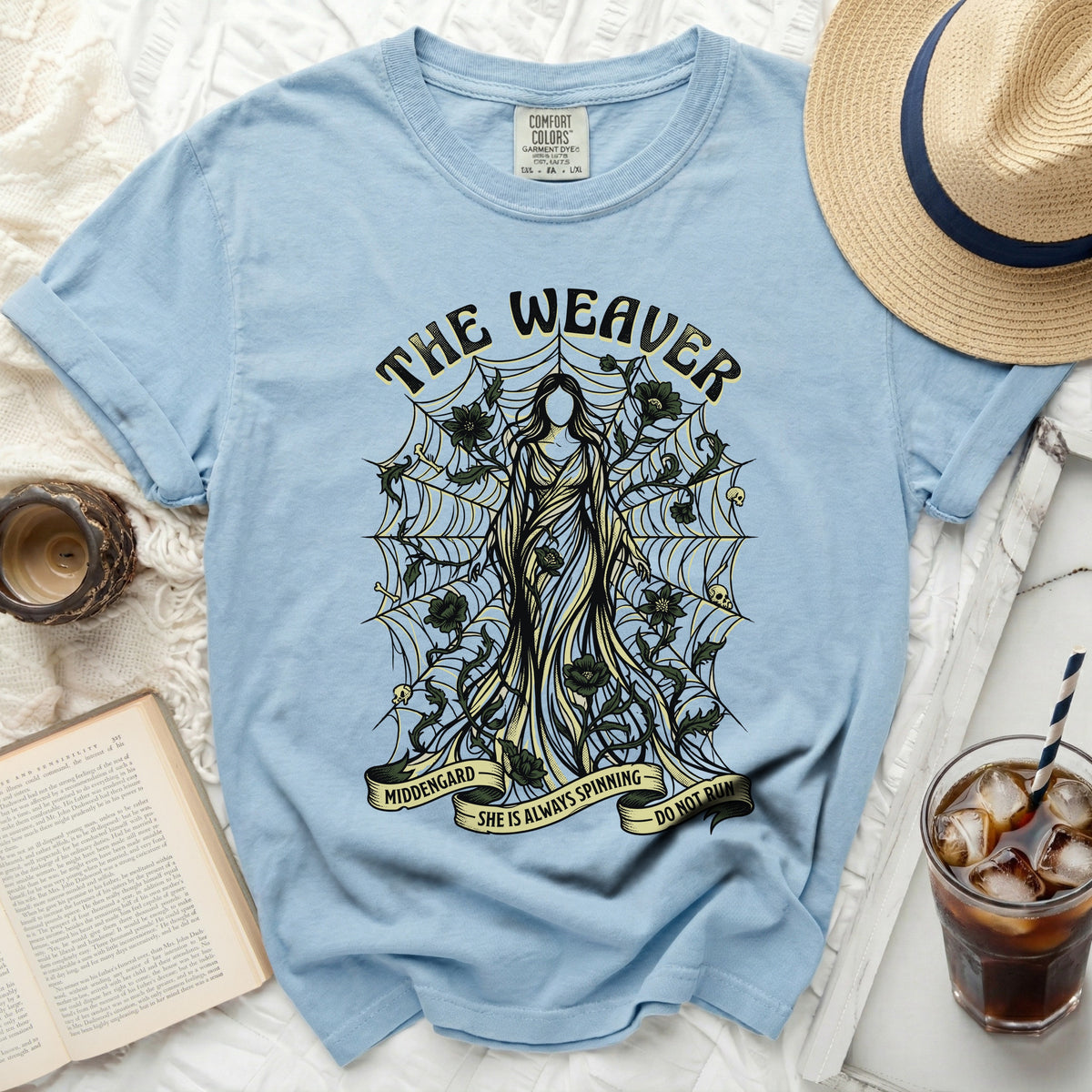Light blue t-shirt with "The Weaver" graphic featuring a figure, spiderwebs, skulls, and flowers.