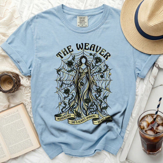 Light blue t-shirt with "The Weaver" graphic featuring a figure, spiderwebs, skulls, and flowers.