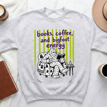 Bigfoot reading a book and drinking coffee on a chair with text "books, coffee, and bigfoot energy".