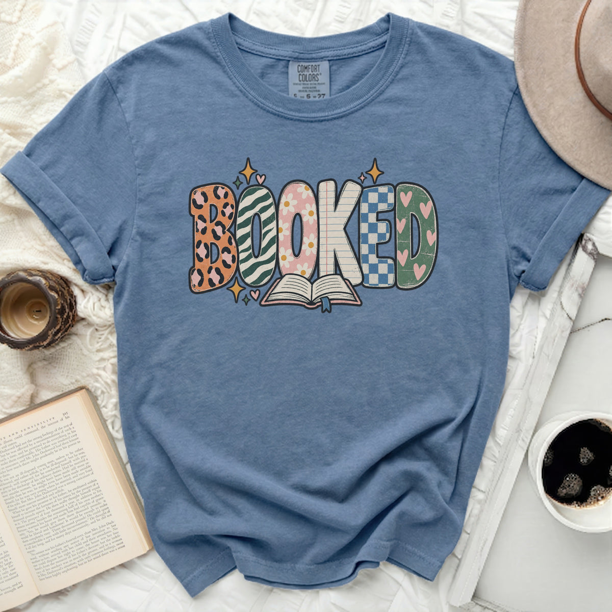 Blue t-shirt with "BOOKED" graphic featuring various patterns and an open book.