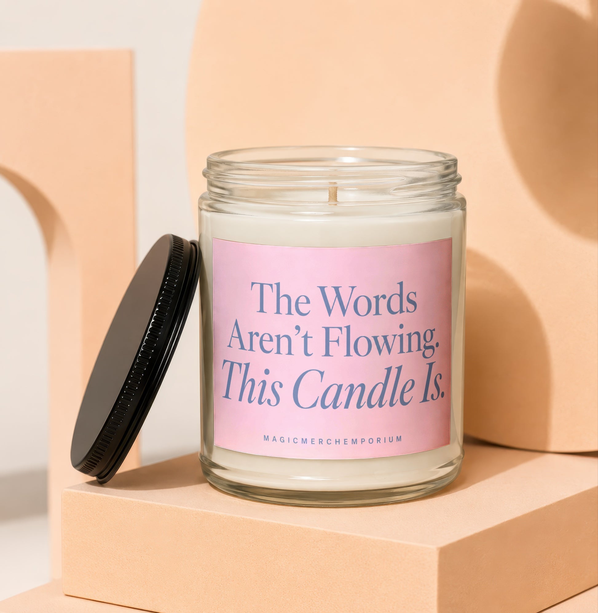 Glass candle with pink label reading "The Words Aren't Flowing. This Candle Is."