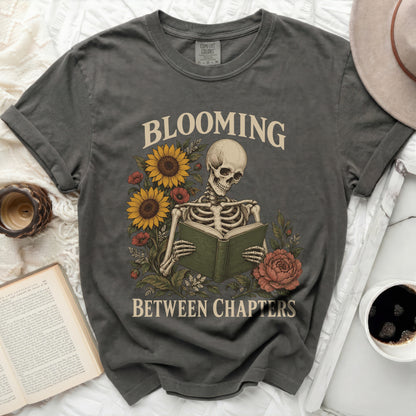 Skeleton reading book with sunflowers and roses, text "BLOOMING BETWEEN CHAPTERS".