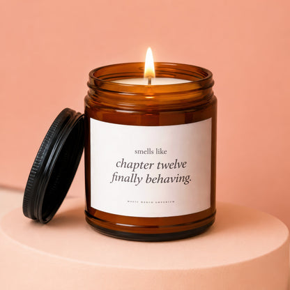 Amber jar candle with lit wick, lid off, labeled "smells like chapter twelve finally behaving."