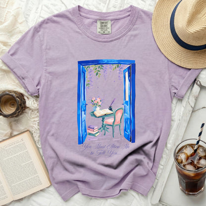 Lavender t-shirt with a graphic of an open blue door with wisteria, a table, chair, books, and quill.