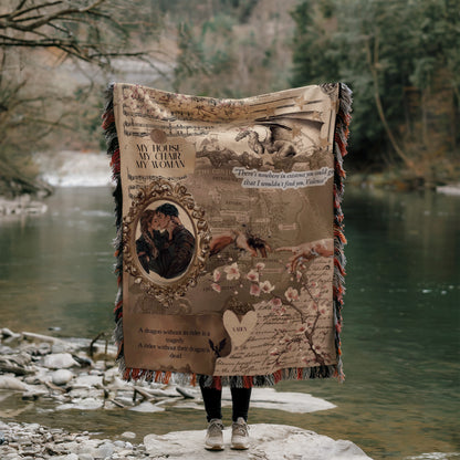 A person stands on a rock, holding a large tapestry or blanket with a dragon and map design, in front of a body of water.