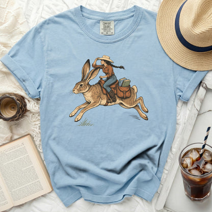 Girl riding a rabbit with books in a saddlebag on light blue t-shirt.