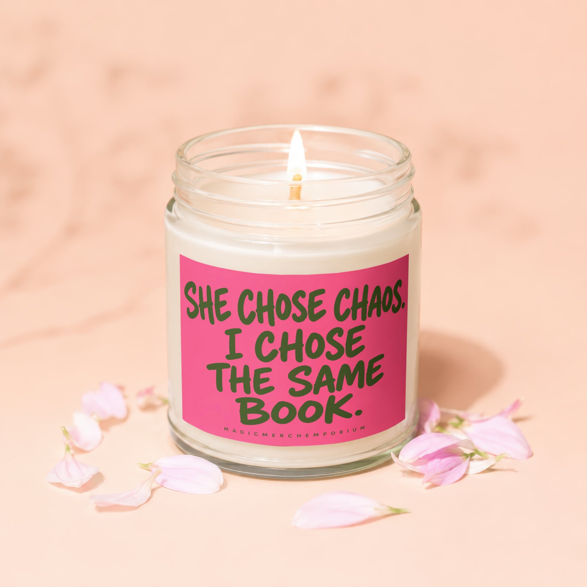 Lit candle in glass jar with pink label reading "SHE CHOSE CHAOS. I CHOSE THE SAME BOOK."