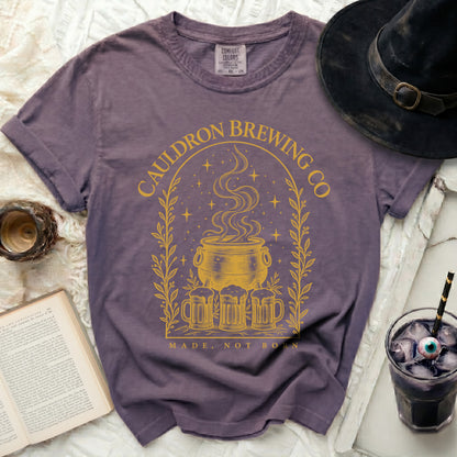 Faded purple crewneck t-shirt featuring a yellow graphic of a cauldron and text reading Cauldron Brewing Co Made Not Born.