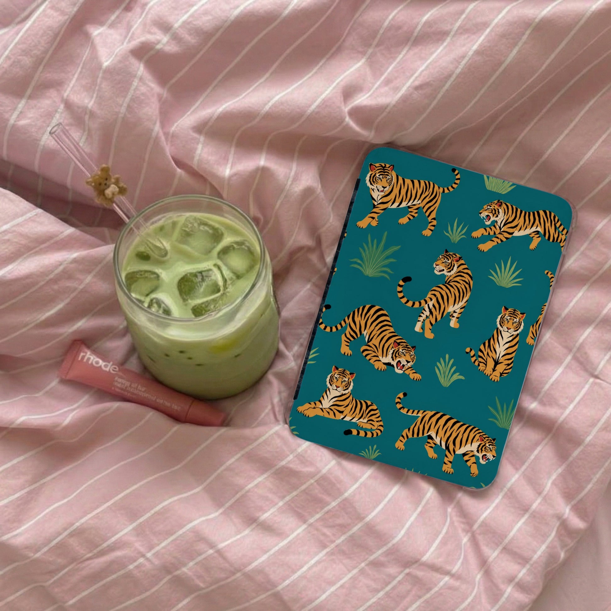 Teal case with tiger pattern next to iced matcha drink on pink striped bedding.