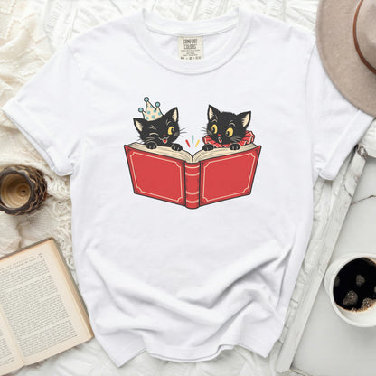 Two black cats read a red book, one wearing a party hat.