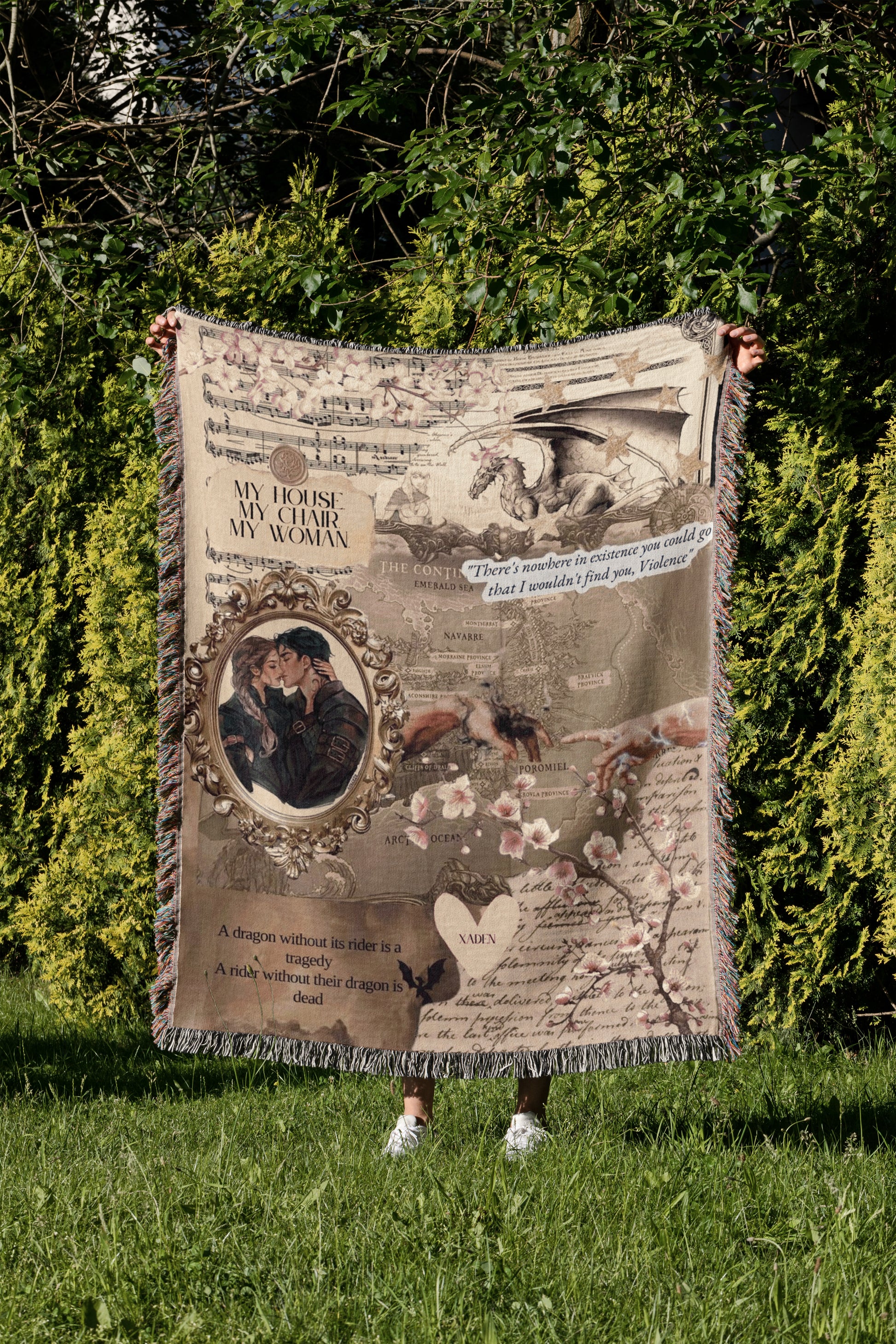 A person is holding a large, ornate blanket with various images and text printed on it, standing in a grassy area with trees in the background.