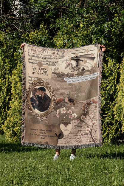 A person is holding a large, ornate blanket with various images and text printed on it, standing in a grassy area with trees in the background.