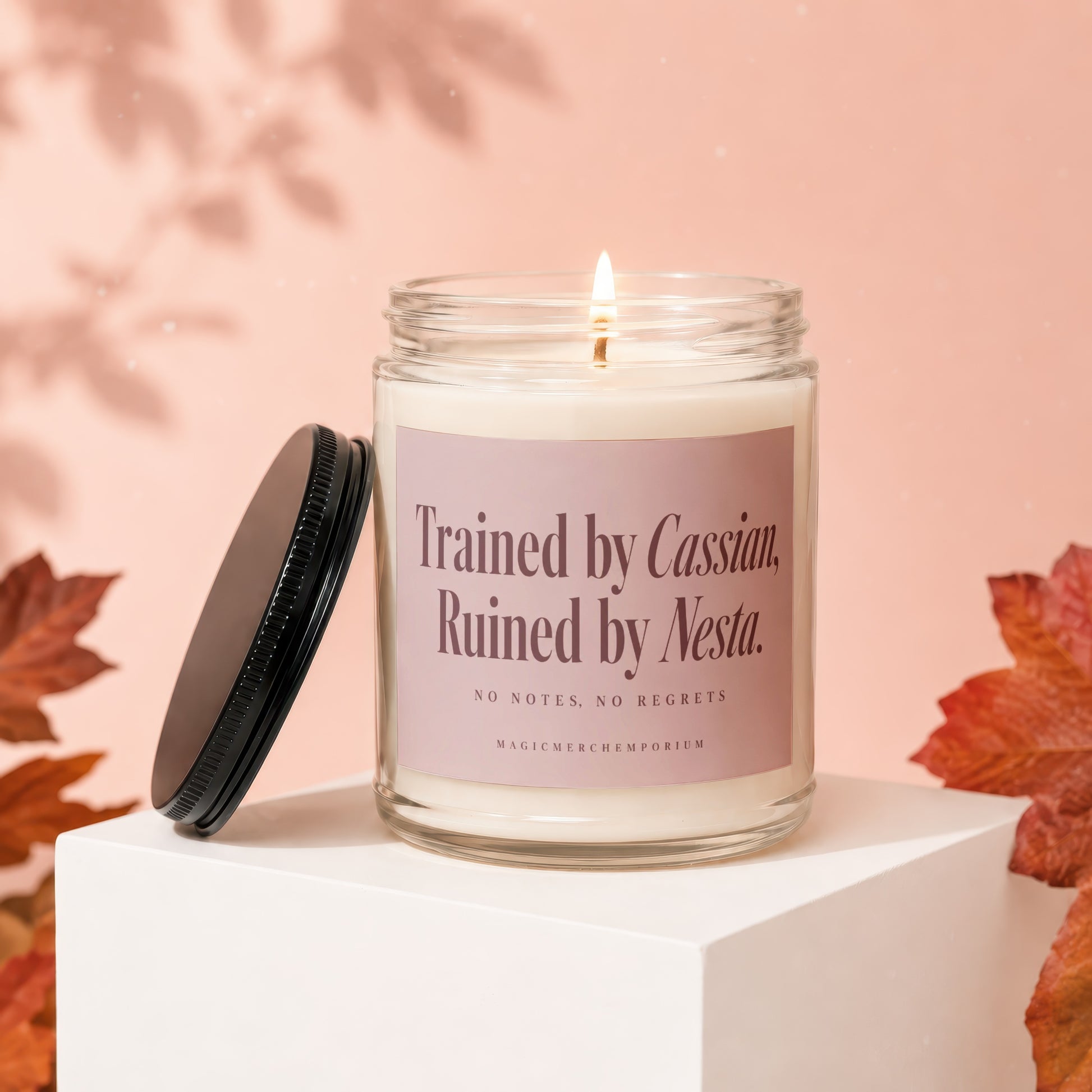 A lit candle in a glass jar with a black lid and pink label resting on a white stand against a pink background with autumn leaves.