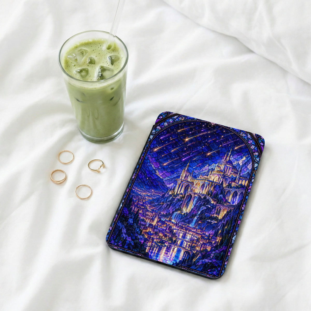 An e-reader with a stained-glass castle case rests on white bedding next to four gold rings and an iced matcha latte.