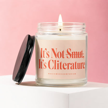 Scented candle in a glass jar with a lit wick and a black lid, reads "It's Not Smut, It's Cliterature".