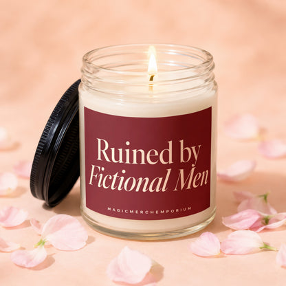 Lit candle with maroon label reading "Ruined by Fictional Men" on textured background with petals.