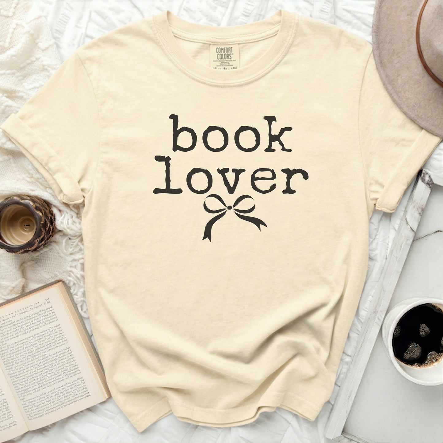 Cream t-shirt with "book lover" and bow graphic, shown with a book and hat.