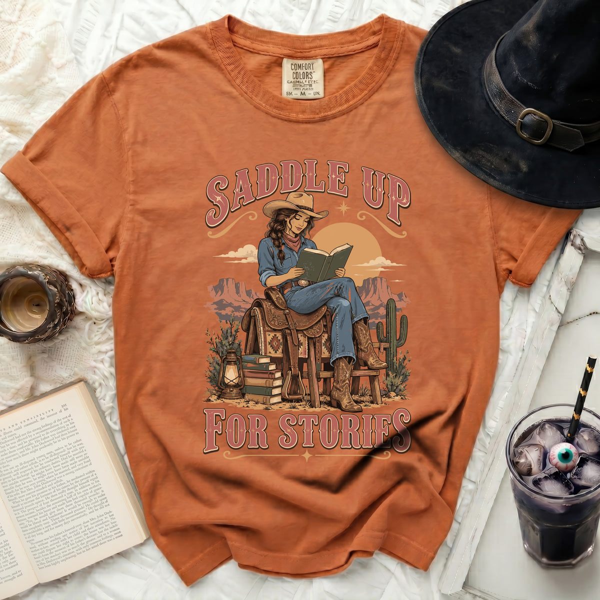 Orange t-shirt with a cowgirl reading a book on a saddle, "Saddle Up For Stories" text.