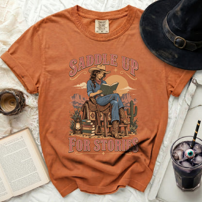 Orange t-shirt with a cowgirl reading a book on a saddle, "Saddle Up For Stories" text.