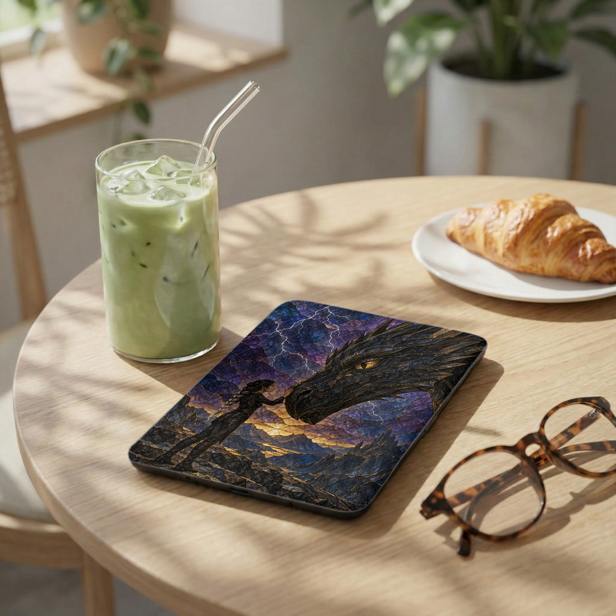 Tablet with dragon and girl artwork, glass of matcha latte, croissant, and glasses.