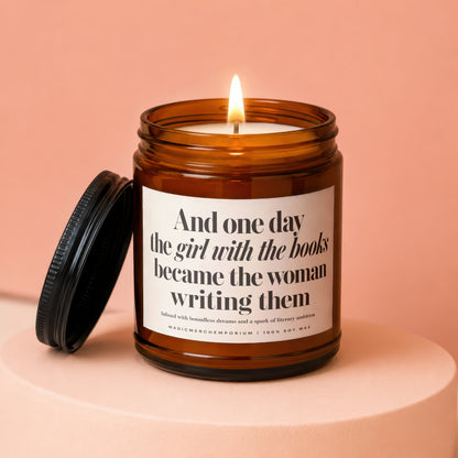 Amber jar candle with lit flame and text "And one day the girl with the books became the woman writing them."