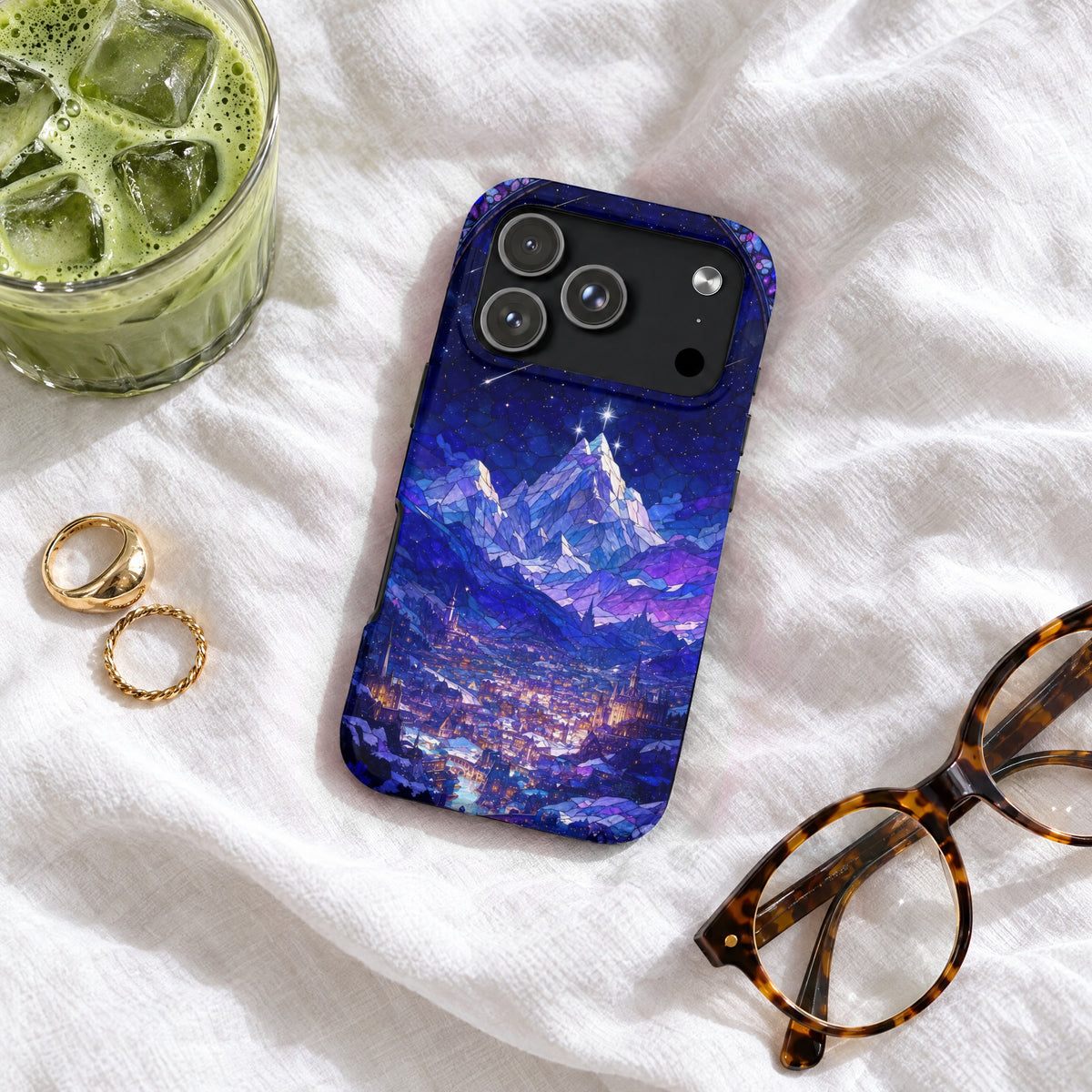 Blue phone case featuring a stained-glass style mountain and city landscape rests on white fabric with glasses, rings, and a drink.
