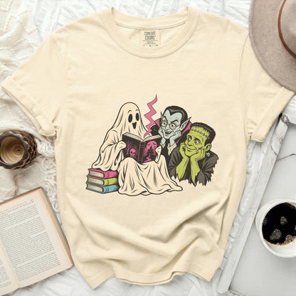 A beige Comfort Colors t-shirt features a graphic of a ghost, vampire, and Frankenstein reading a book.