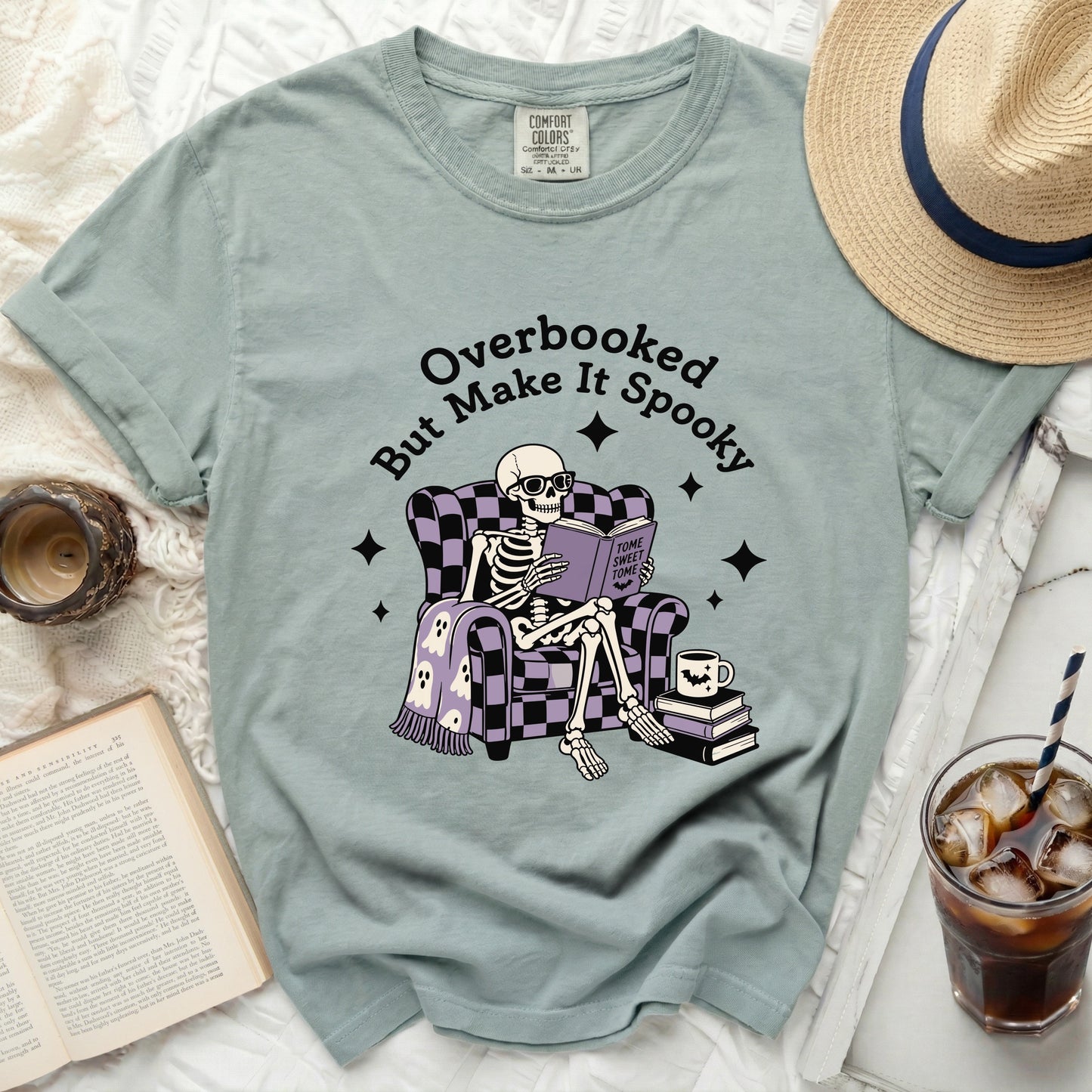 Skeleton in chair reading book on "Overbooked But Make It Spooky" t-shirt.