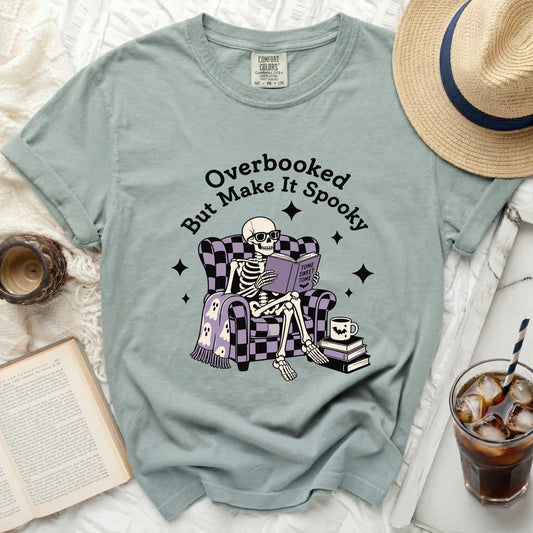 Skeleton in chair reading book on "Overbooked But Make It Spooky" t-shirt.
