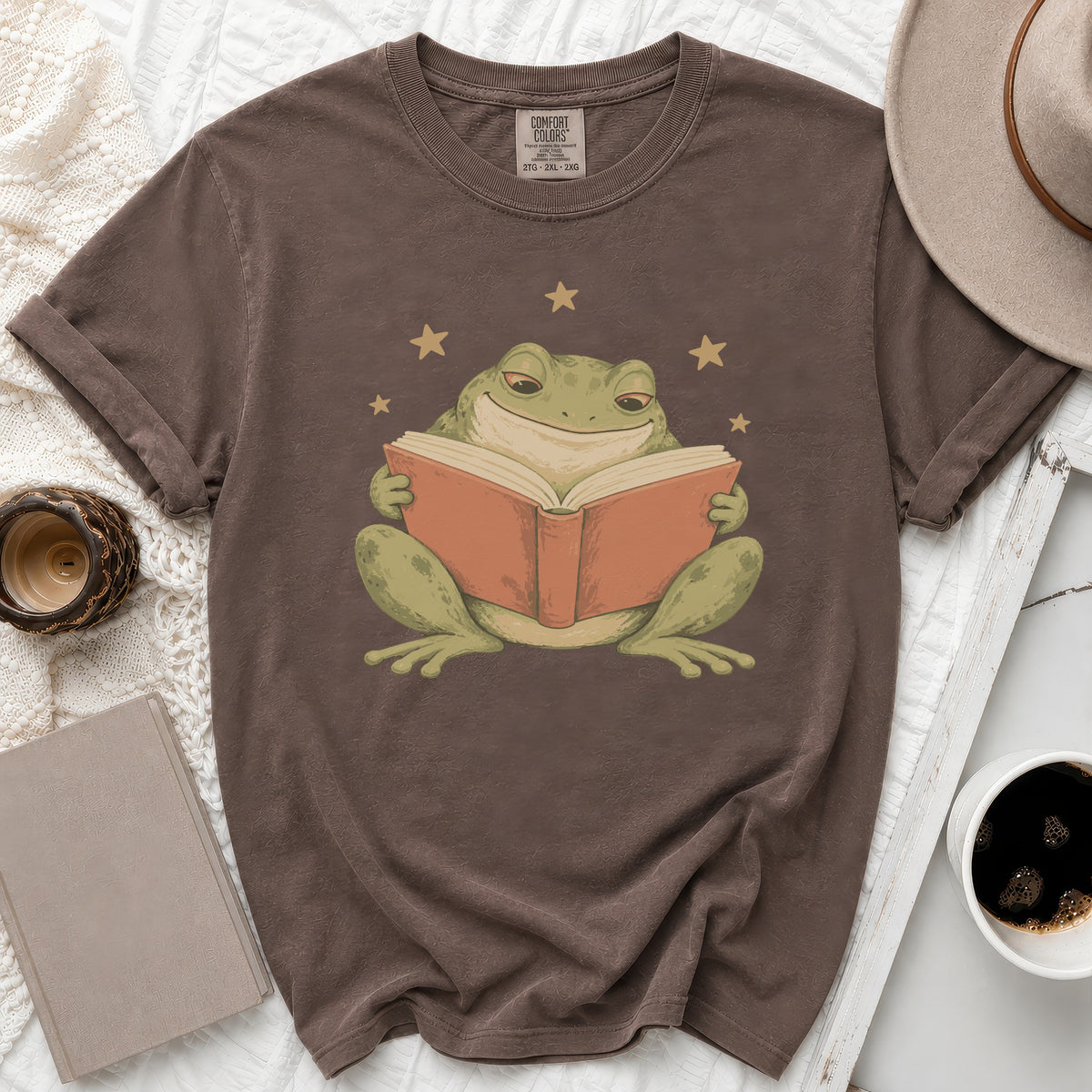 Brown t-shirt with graphic of a smiling frog reading a book surrounded by stars.