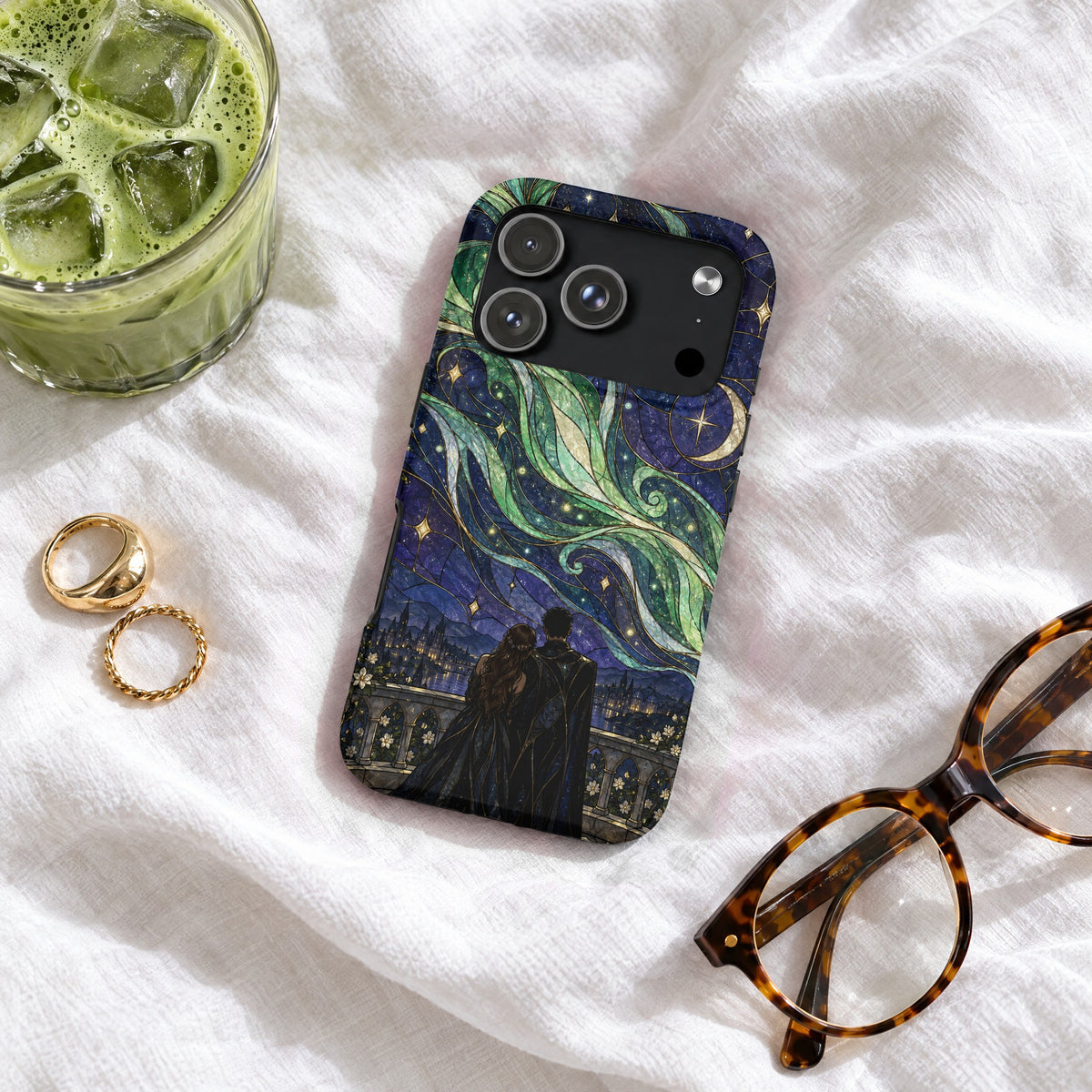 An iPhone case featuring a stained glass-style night sky illustration lies on wrinkled white fabric with accessories.