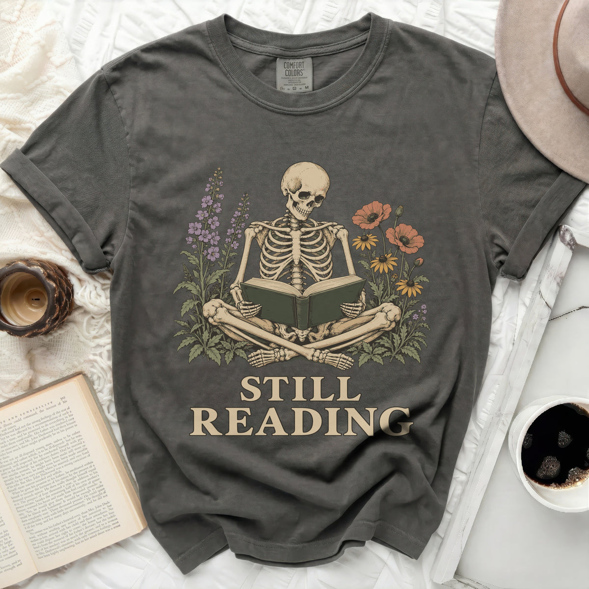 Skeleton reading a book surrounded by flowers, text reads "STILL READING".