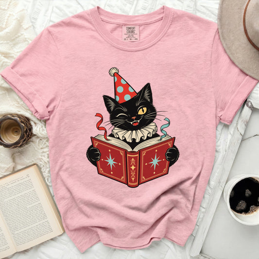 Pink t-shirt with graphic of winking black cat in party hat reading a book.