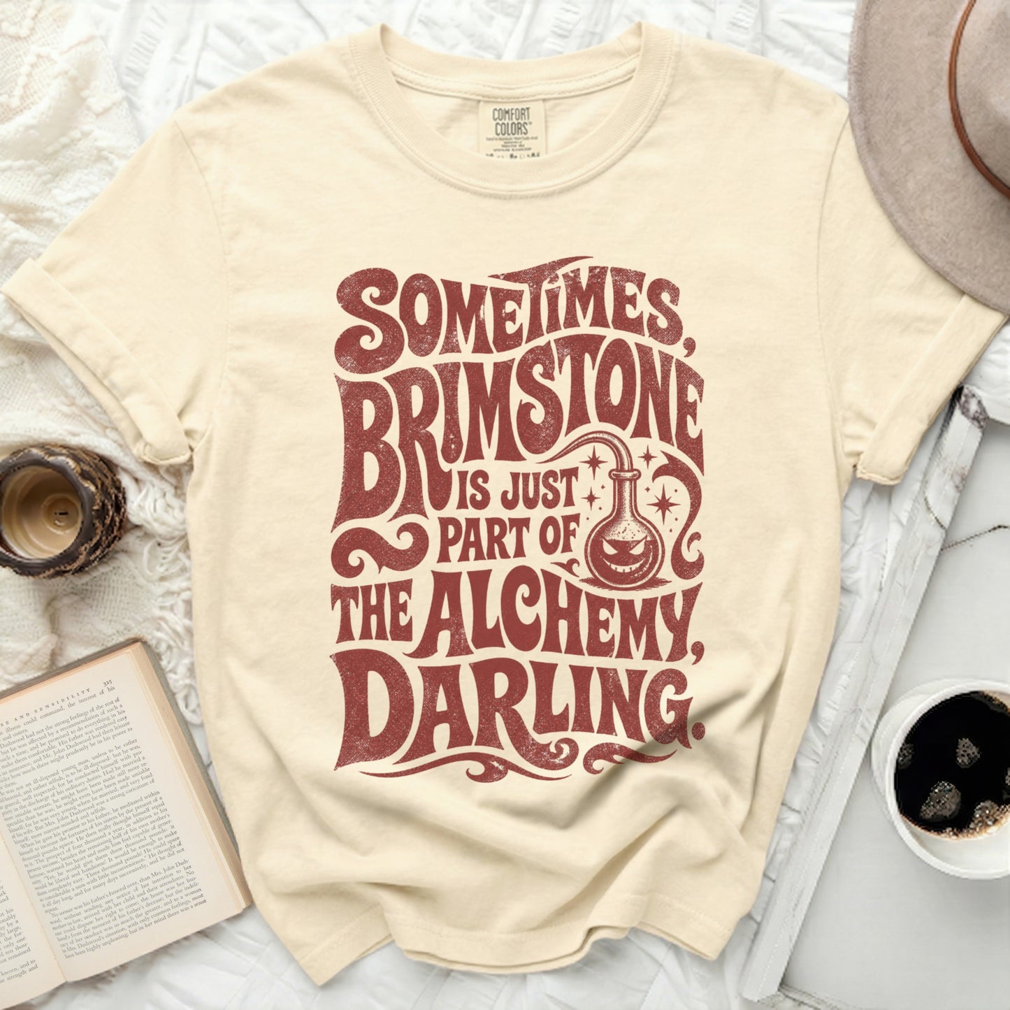 Cream t-shirt with "Sometimes Brimstone is Just Part of the Alchemy, Darling" text.